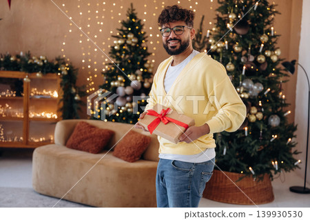 Gift box is in hands. Man posing in front of a Christmas tree in a festive home setting 139930530