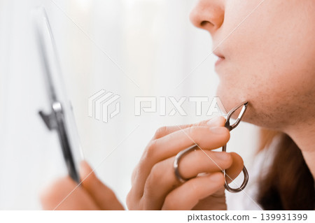 A woman looks in a mirror during her home beauty routine, using a tool for facial hair removal to pluck unwanted chin hair caused by hormonal changes. A woman looks in a mirror during her home beauty routine, using a tool for facial hair removal to pluck unwanted chin hair caused by hormonal changes. 139931399
