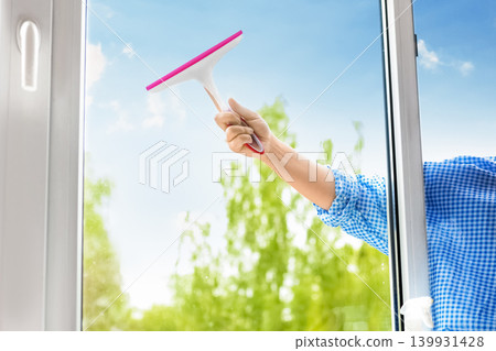 Close-up of a hand washing a window with a squeegee against a bright sunny blue sky and green trees. Ideal for spring cleaning and house chores concepts. Close-up of a hand washing a window with a squeegee against a bright sunny blue sky and green trees. Ideal for spring cleaning and house chores concepts. 139931428