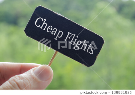 Hand holding a dark paper label showing cheap flights against a blurred outdoor background. Hand holding a dark paper label showing cheap flights against a blurred outdoor background. 139931985