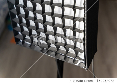 Black and white softbox lighting equipment with a grid pattern, positioned on a stand, showcasing a textured surface and a blurred background of a studio environment Black and white softbox lighting equipment with a grid pattern, positioned on a stand, showcasing a textured surface and a blurred background of a studio environment 139932078