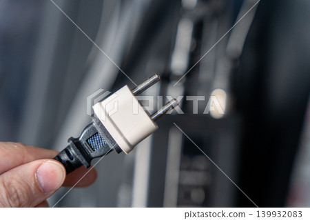 Hand holding a two-prong electrical plug with a black cord, positioned near an electrical outlet in a modern indoor setting Hand holding a two-prong electrical plug with a black cord, positioned near an electrical outlet in a modern indoor setting 139932083