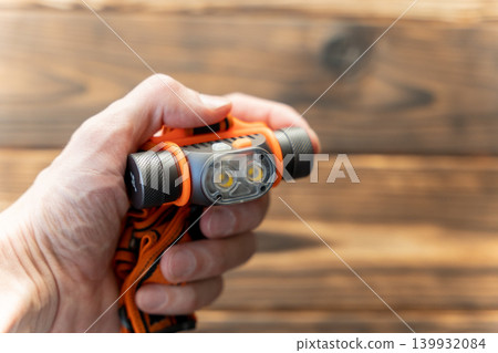 Hand holding an orange and gray handheld flashlight with dual LED lights against a wooden background, showcasing its design and functionality for outdoor use 139932084