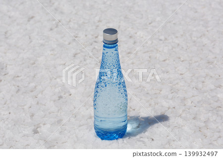 Blue glass water bottle standing on a textured white salt crust. Concept of absolute purity, refreshing hydration and natural mineral source. Blue glass water bottle standing on a textured white salt crust. Concept of absolute purity, refreshing hydration and natural mineral source. 139932497