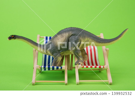 Funny toy sauropod dinosaur trying to fit on two miniature beach deck chairs, green background. 139932532