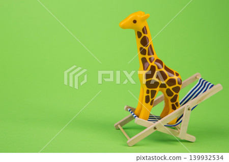 Funny toy giraffe standing on a miniature beach deck chair against a green background. Funny toy giraffe standing on a miniature beach deck chair against a green background. 139932534