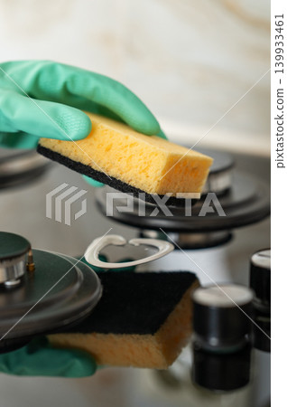 A close-up of a person's hand in a rubber glove cleaning a gas cooktop with a sponge. A close-up of a person's hand in a rubber glove cleaning a gas cooktop with a sponge. 139933461