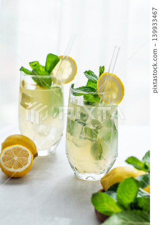 A summer drink with lemon, mint, and ice in a two glasses against a sunny window background. 139933467