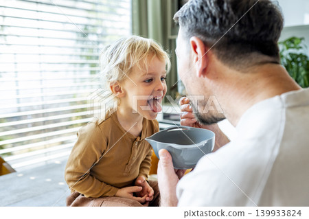 Dad feeding toddler during family mealtime in modern kitchen. Dad feeding toddler during family mealtime in modern kitchen. 139933824