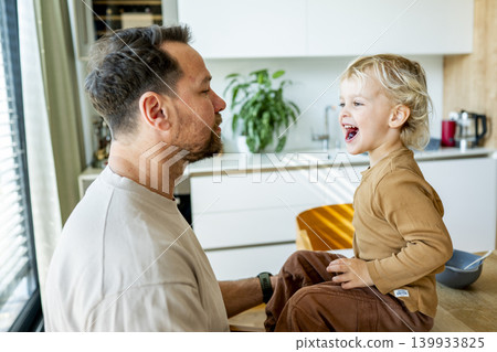 Dad feeding toddler during family mealtime in modern kitchen. Dad feeding toddler during family mealtime in modern kitchen. 139933825