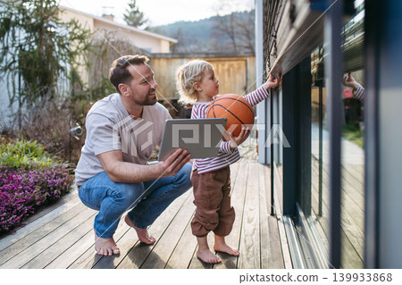 Father controlling smart home exterior blinds using tablet while standing with toddler son. Father controlling smart home exterior blinds using tablet while standing with toddler son. 139933868