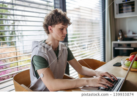 Teen boy with diabetes working on school project on laptop at kitchen table. 139933874
