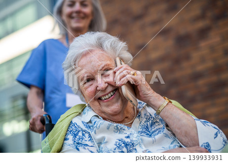 Older nurse pushing senior woman in wheelchair while making phone call. Older nurse pushing senior woman in wheelchair while making phone call. 139933931
