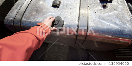 Hand of a person in an orange jacket opening the fuel cap on a metallic gas tank of a truck, showcasing the tank's surface and surrounding details Hand of a person in an orange jacket opening the fuel cap on a metallic gas tank of a truck, showcasing the tank's surface and surrounding details 139934060