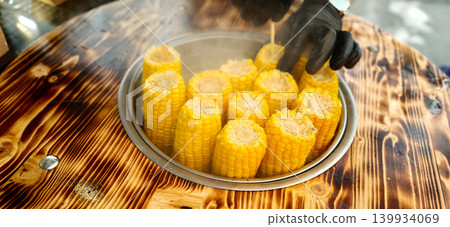 Boiled corn on a wooden table, vendor wearing black gloves prepares fresh corn cobs for sale in a street food setting, steam rising from the hot food Boiled corn on a wooden table, vendor wearing black gloves prepares fresh corn cobs for sale in a street food setting, steam rising from the hot food 139934069