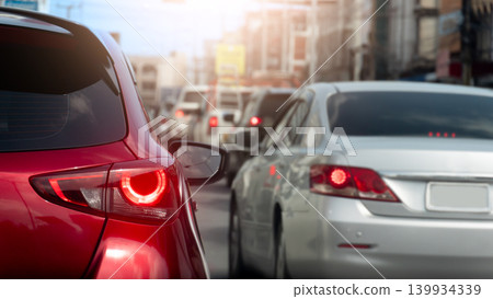 Shallow depth of field shot focusing on the rear side of a glossy red car stuck in traffic. Silver sedan and a long line of cars stretch against the sunlit city backdrop. Shallow depth of field shot focusing on the rear side of a glossy red car stuck in traffic. Silver sedan and a long line of cars stretch against the sunlit city backdrop. 139934339