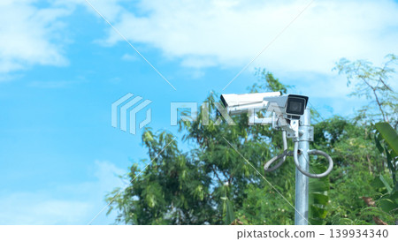 White outdoor security CCTV camera mounted on a metal pole against a vibrant blue sky with light clouds. Lush green tree branches and foliage are visible in the background frame. White outdoor security CCTV camera mounted on a metal pole against a vibrant blue sky with light clouds. Lush green tree branches and foliage are visible in the background frame. 139934340