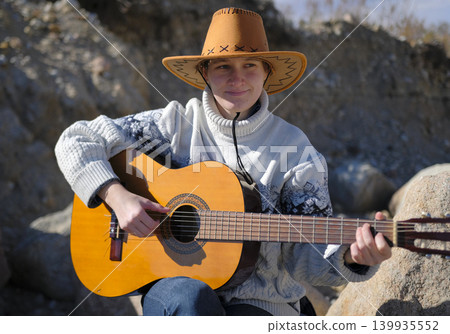An authentic portrait of a female guitarist. A young woman in a cowboy hat and a white sweater is playing guitar in a rocky gorge. She smiles and enjoys what is happening An authentic portrait of a female guitarist. A young woman in a cowboy hat and a white sweater is playing guitar in a rocky gorge. She smiles and enjoys what is happening 139935552