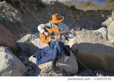 A female cowboy plays guitar sitting on a large rock surrounded by other boulders. A musician learns to play the guitar on a sunny day on a pile of stones. The concept of freedom and adventure A female cowboy plays guitar sitting on a large rock surrounded by other boulders. A musician learns to play the guitar on a sunny day on a pile of stones. The concept of freedom and adventure 139935556