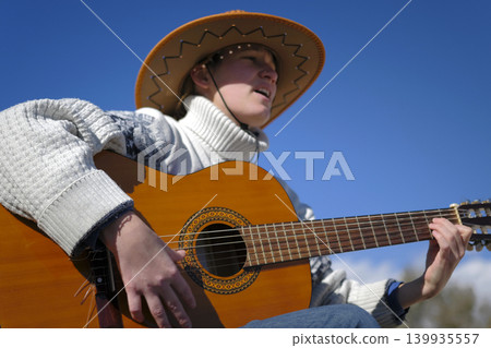 Portrait of a female guitarist singing songs with a guitar against a blue sky background. A musician in a cowboy hat and a white sweater plays the guitar, focusing on the hand and strings Portrait of a female guitarist singing songs with a guitar against a blue sky background. A musician in a cowboy hat and a white sweater plays the guitar, focusing on the hand and strings 139935557