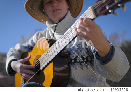 Guitar lessons. A white young female student in a cowboy hat and a white sweater is playing guitar on the street. Focus on the guitar strings. The concept of relaxation and leisure 139935558
