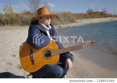 A lonely female guitarist looks into the distance by the sea and dreams of a better life. A sad musician in a cowboy hat is sitting on an empty beach with a guitar in his hands 139935569
