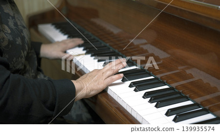 An elderly woman plays the piano, close-up on her hands. Focus on the old man's hand pressing the piano keys. Music school or concert. The piano is a large wooden instrument An elderly woman plays the piano, close-up on her hands. Focus on the old man's hand pressing the piano keys. Music school or concert. The piano is a large wooden instrument 139935574