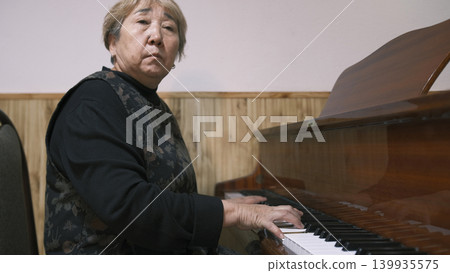 An old elderly Asian woman is sitting at the piano and playing a melody. A strict teacher at a music school. Piano lessons. a classical musical instrument An old elderly Asian woman is sitting at the piano and playing a melody. A strict teacher at a music school. Piano lessons. a classical musical instrument 139935575