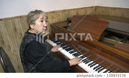 An elderly Asian woman plays the piano in a music school classroom. She's focused on her game. Piano lessons. An old pianist at work. A musician An elderly Asian woman plays the piano in a music school classroom. She's focused on her game. Piano lessons. An old pianist at work. A musician 139935576