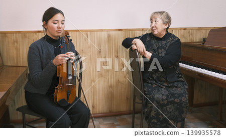 A lesson at a music school. An elderly Asian female teacher looks at her young student. The student is sitting with a violin in his hands, and the teacher is at the piano A lesson at a music school. An elderly Asian female teacher looks at her young student. The student is sitting with a violin in his hands, and the teacher is at the piano 139935578