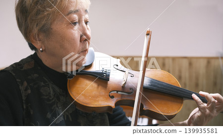 An old Asian woman plays the violin, close-up. Violin lessons. An elderly teacher shows how to play the violin correctly. A focused musician An old Asian woman plays the violin, close-up. Violin lessons. An elderly teacher shows how to play the violin correctly. A focused musician 139935581