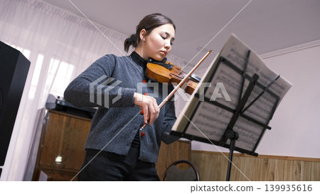 An Asian teenage student is learning to play the violin. A young woman looks at the sheet music and plays the violin at a music school. The musician is focused on the rehearsal An Asian teenage student is learning to play the violin. A young woman looks at the sheet music and plays the violin at a music school. The musician is focused on the rehearsal 139935616