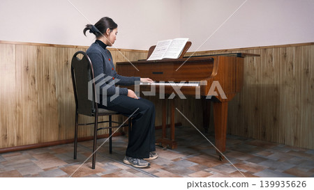 A young Asian student is learning to play the piano. Music school. A woman is sitting at the piano with a music book in front of her. She plays the piano and is focused on music 139935626