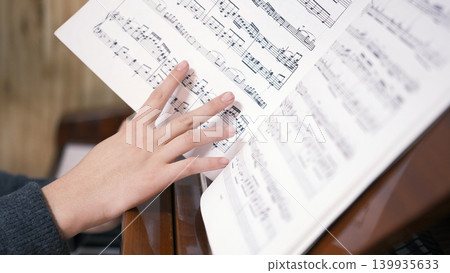 Close-up of a young woman turning the pages of a music book. A musician plays the piano, holding notes in front of him. A piano student is learning a new tune Close-up of a young woman turning the pages of a music book. A musician plays the piano, holding notes in front of him. A piano student is learning a new tune 139935633