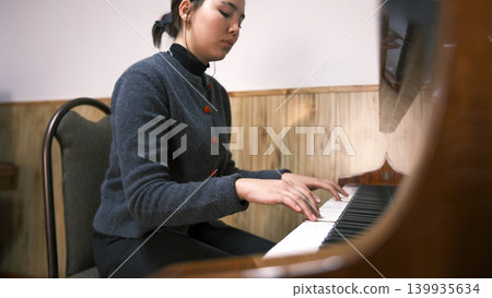 A music school student plays the piano. A young Asian woman is learning to play the piano. She is wearing a grey sweater and black trousers. The pianist is focused on her playing A music school student plays the piano. A young Asian woman is learning to play the piano. She is wearing a grey sweater and black trousers. The pianist is focused on her playing 139935634