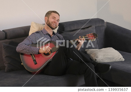 A cute, pretty guitarist plays the guitar. A smiling man is sitting on the couch and composing music on the guitar. Guitar lessons. The scene is relaxed and happy 139935637