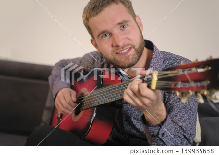 A white unshaven man is sitting on the couch with a guitar in his hands. He smiles and sings songs to the guitar. Guitar lessons. Focus on the guitarist's face 139935638
