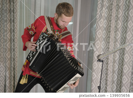 A guy in a red national shirt holds a large accordion in his hands. The musician plays the accordion, opening the bellows of the accordion wide. Accordion playing lessons A guy in a red national shirt holds a large accordion in his hands. The musician plays the accordion, opening the bellows of the accordion wide. Accordion playing lessons 139935656