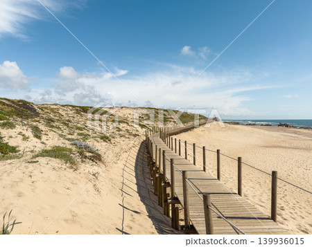 A winding wooden boardwalk stretches across golden sand dunes toward the blue Atlantic Ocean under a bright sky at Passadicos da Ribeira do Espirito Santo in Miramar A winding wooden boardwalk stretches across golden sand dunes toward the blue Atlantic Ocean under a bright sky at Passadicos da Ribeira do Espirito Santo in Miramar 139936015
