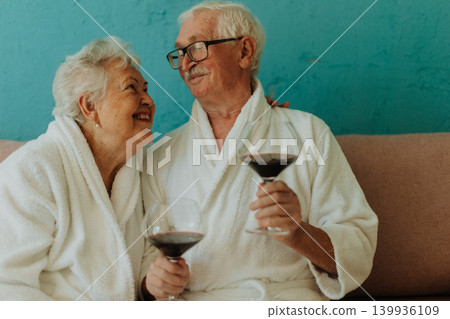 Happy senior couple sitting together in bathrobe on sofa with glass of wine. Happy senior couple sitting together in bathrobe on sofa with glass of wine. 139936109