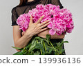 Close-up of female hands hugging a large armful bouquet of fresh pink peonies against a grey background. Variety Sarah Bernhardt, Alexander Fleming, Kansas. Paeonia lactiflora. 139936136