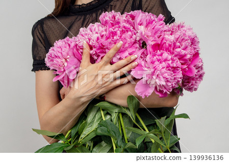 Close-up of female hands hugging a large armful bouquet of fresh pink peonies against a grey background. Variety Sarah Bernhardt, Alexander Fleming, Kansas. Paeonia lactiflora. Close-up of female hands hugging a large armful bouquet of fresh pink peonies against a grey background. Variety Sarah Bernhardt, Alexander Fleming, Kansas. Paeonia lactiflora. 139936136