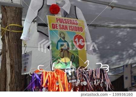San isidro festival bracelets and devotional medals displayed at market stall with traditional costume and promotional sign for religious celebration souvenirs San isidro festival bracelets and devotional medals displayed at market stall with traditional costume and promotional sign for religious celebration souvenirs 139936160