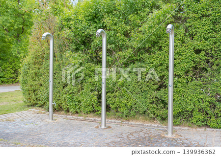 Row of three modern stainless steel outdoor showers installed in a public resort park. Convenient infrastructure against a lush green hedge backdrop 139936362
