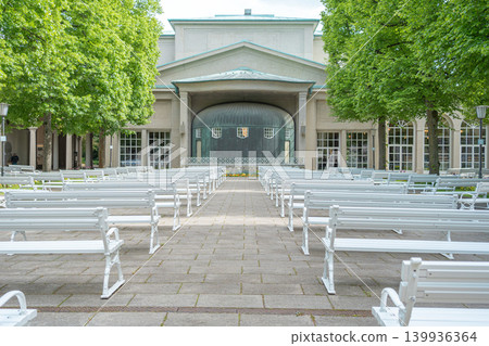 Symmetric view of empty white wooden benches arranged for an outdoor concert in front of the music pavilion. Green trees and pavement in a quiet spa resort Symmetric view of empty white wooden benches arranged for an outdoor concert in front of the music pavilion. Green trees and pavement in a quiet spa resort 139936364
