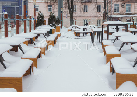 Tables and benches covered with snow stand on the city street on a pavement next to the restaurant winter. Tables and benches covered with snow stand on the city street on a pavement next to the restaurant winter. 139936535