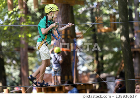 Young child boy in safety harness and helmet attached with carbine to cable on rope way in park. Young child boy in safety harness and helmet attached with carbine to cable on rope way in park. 139936637