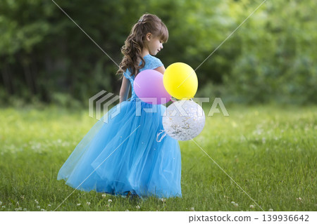 Profile full-length portrait of pretty little blond long-haired girl in long blue dress with colorful balloons standing in blooming field on blurred green trees background. Happy childhood concept. Profile full-length portrait of pretty little blond long-haired girl in long blue dress with colorful balloons standing in blooming field on blurred green trees background. Happy childhood concept. 139936642