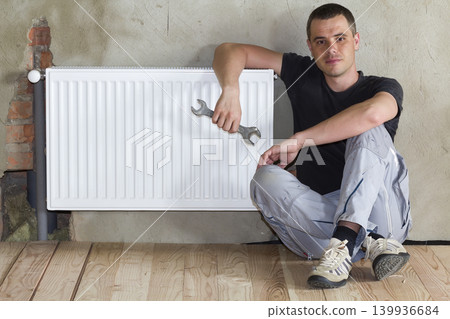 Young handsome plumber sitting on floor with wrench in hand near successfully installed heating radiator in empty room of a newly built apartment or house. Construction, maintenance and repair concept 139936684
