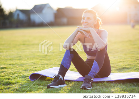 Young sportive woman in sports clothes sitting on training mat before doing exercises in field at sunrise. Young sportive woman in sports clothes sitting on training mat before doing exercises in field at sunrise. 139936734